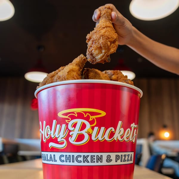 A hand pulls a crispy fried chicken drumstick from a red Holy Buckets bucket filled with chicken.