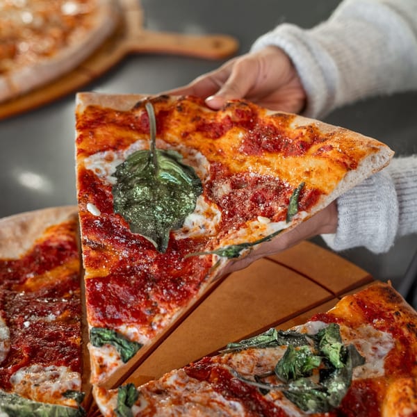 A hand in a grey sweater lifts a slice of pizza with red sauce, cheese, and basil.