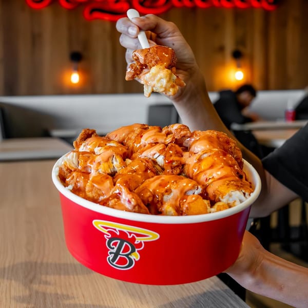 A hand holds a Holy Buckets Halal Chicken & Chicken bucket filled with saucy fried chicken and mac and cheese.